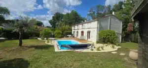 Maison traditionnelle avec volets blancs, piscine, et grande terrasse en bois, entourée d'un jardin luxuriant sous un ciel bleu d'été.