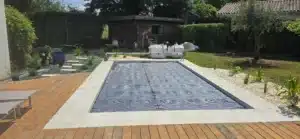 Aménagement paysager d'une piscine avec terrasse bois et margelles claires. Des ouvriers plantent des végétaux et préparent les massifs de galets.