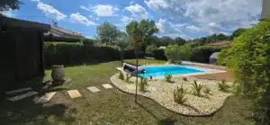 Jardin ensoleillé, piscine creusée d'un bleu intense bordée de bois et de gravier décoratif. Haies de confidentialité et ciel nuageux.