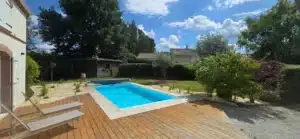 Vue sur une piscine ensoleillée bordée d'une terrasse en bois et de margelles claires. Jardin luxuriant avec graviers et chaises longues.