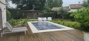 Grande terrasse en bois brun et piscine couverte, entourée de dalles claires. Aménagement de jardin en cours avec plantes, cabane et outils.