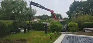 Trois paysagistes transplantent un grand olivier en pot dans un jardin résidentiel, manœuvré par une grue Palfinger près de la piscine.